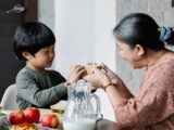 Side view of content Asian boy taking homemade sandwich from grandmother while sitting near served table during breakfast time in kitchen