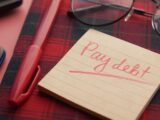 a note that says pay debt next to a pen and glasses