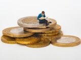 Miniature person sitting on stack of coins reading newspaper