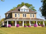A tan two-story farmhouse with a wrap-around porch