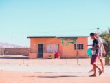 a woman walking across a dirt field next to a building