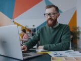 A man with a red beard and glasses using a laptop in a workspace